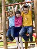 Three children hang playfully from playground bars. Two boys, on either side, wear blue and yellow shirts. A girl in a pink shirt hangs in the middle. They smile happily, enjoying their time outdoors.
