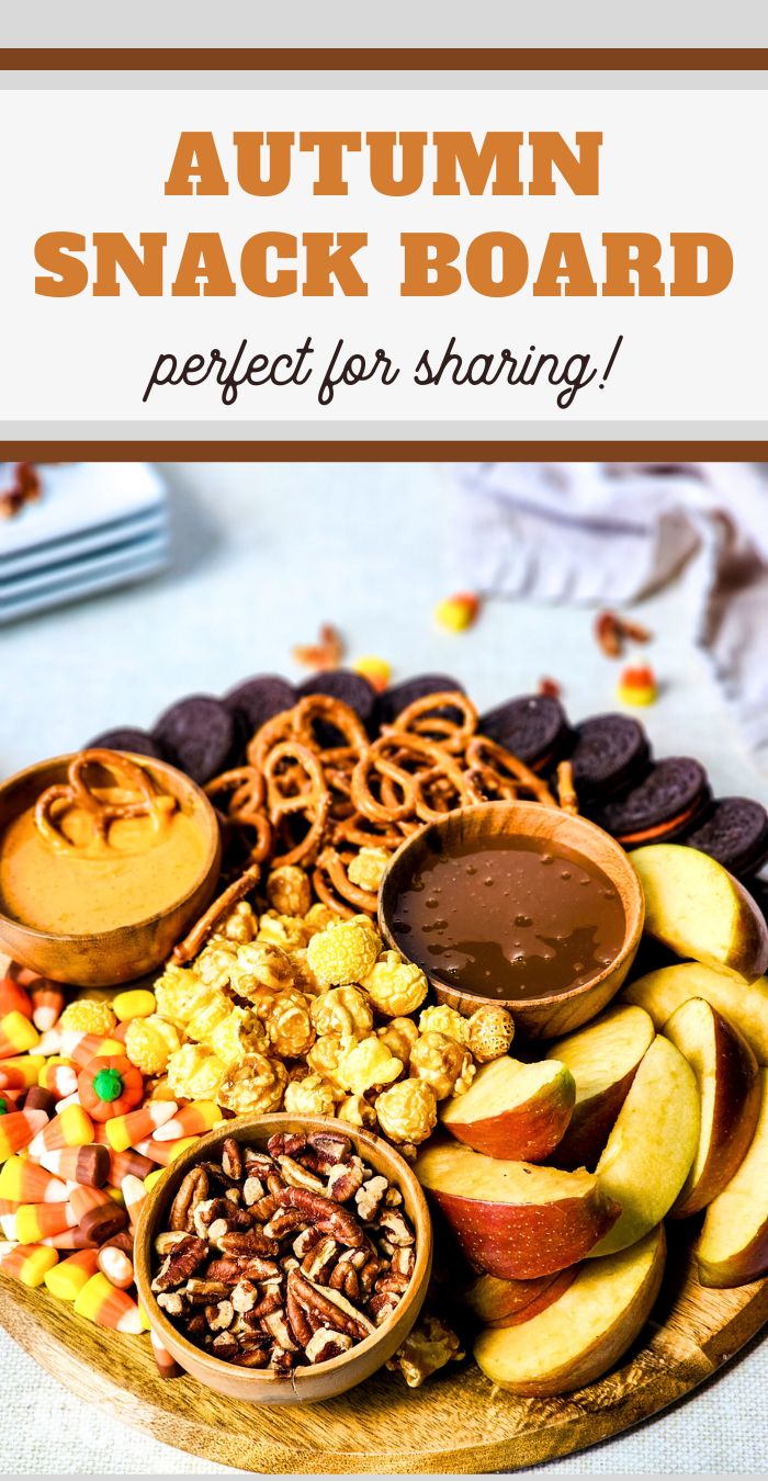 A round wooden board filled with autumn-themed snacks, including apple slices, pretzels, pecans, candy corn, popcorn, and chocolate cookies. Small bowls of caramel and peanut butter dips are placed on the board. Discover this Autumn Snack Board Recipe&mdash;perfect for sharing!.